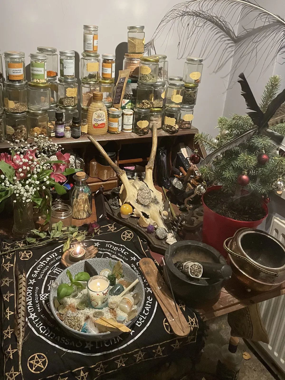 Jasmine's altar at night: a lit candle and offering bowl on the wheel-of-the-year cloth, with antlers, fir and the herb jars behind
