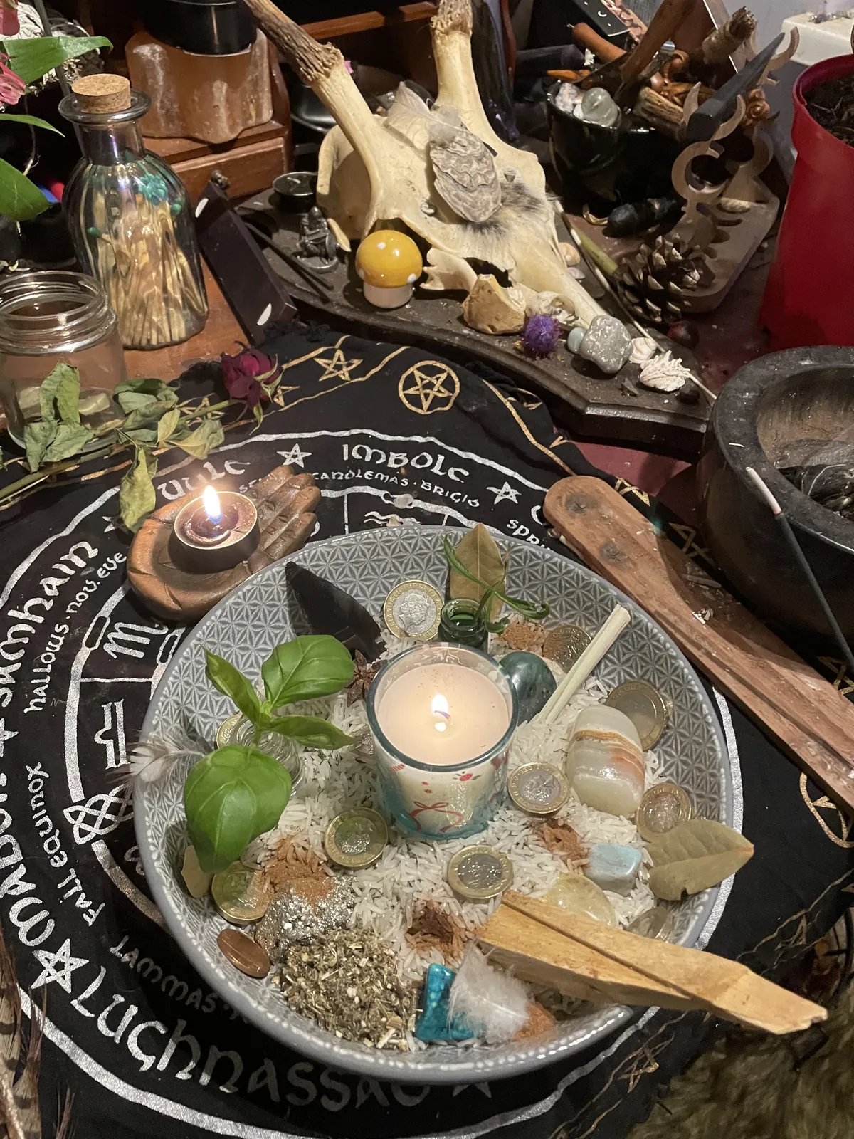 An offering bowl on Jasmine's altar: a lit candle set in rice with coins, basil leaves and crystals, on the wheel-of-the-year cloth