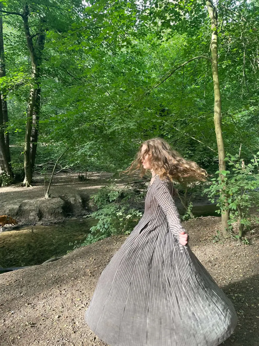 Jasmine turning in a striped dress beside a woodland stream, hair caught in the light